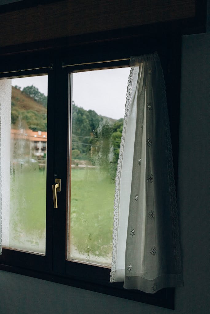 Peaceful view of the countryside through a rustic window with lace curtains and condensation.